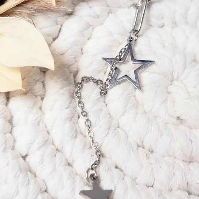 Collier étoile argenté en acier inoxydable, gros maillons et pendentifs étoiles, gros plan sur fond blanc texturé.