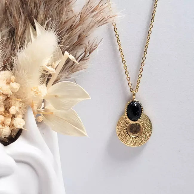 Collier doré avec pendentif médaillon texturé et cabochon bleu nuit pailleté, vu de face près de fleurs séchées, fond blanc.